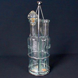 <b>Galheteiro </b>em vidro transparente com suporte em metal cromado, composto por três recipientes cilíndricos. Um deles com tampa original em vidro lapidado e dois sem tampa. Apresenta inscrição em relevo nas garrafas. Alt. 33 cm