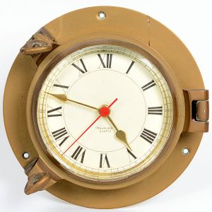<b>Relógio náutico de parede </b>em bronze patinado, com moldura em forma de escotilha, mostrador com algarismos romanos e ponteiro de segundos central em vermelho. Movimento quartz, marca Nauticalia, Inglaterra. Diâm. 23 cm
