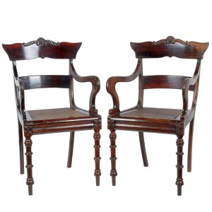 <b>Par de cadeiras de braços </b>em madeira escura entalhada, encosto levemente curvo com friso superior recortado e ornado ao centro, braços em voluta, assento em palhinha e pernas torneadas com anéis sucessivos. Alt. 97 cm