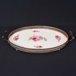Antiga <b>bandeja </b>em metal, com placa em porcelana branca e delicada composição floral em tons de rosa. Estrutura oval com alças laterais e borda rendilhada. Apresenta rachaduras visíveis na porcelana. Compr. 50 cm