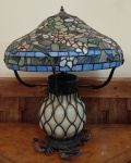<b>Luminária de mesa </b>estilo Tiffany, com cúpula em vitral policromado de motivos florais, corpo globular em vidro leitoso com trama metálica aplicada, sobre  base em metal trabalhado com pés em volutas. Alt. 64 cm