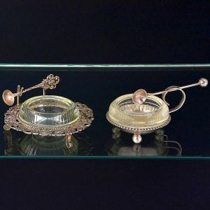 <b>Dois saleiros </b>em metal com recipientes em vidro, acompanhados de colheres. Bases ornamentadas apoiadas sobre pés esféricos, um com borda decorada por motivo floral, enquanto o outro segue desenho geométrico.