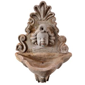 Fonte de parede em pedra esculpida com espaldar ornamentado por volutas e grande concha superior, tendo ao centro máscara de querubim atuando como bica. Pia de borda sinuosa. Séc. XX. Med. 110 x 75 cm