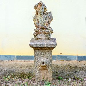 <b>Escultura em pedra </b>entalhada, representando figura masculina jovem trajando vestes de época, apoiada sobre base cilíndrica com orifício frontal que sugere antigo uso como bica ou chafariz. Alt. 130 cm