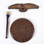 <b>Tambor </b>circular com superfície gravada com motivos que lembram pegadas ou sinais rituais decorativos, com suporte em forma de águia. Acompanha baqueta em madeira com ponta revestida. Med. 42 x 35 cm