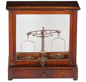 <b>Balança de precisão </b>com estrutura metálica protegida por caixa vitrine em madeira envernizada com faces envidraçadas. Base com gaveta frontal para acondicionamento de pesos. Med. 37 x 35 x 17 cm