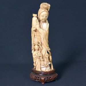 <b>Estatueta chinesa </b>em marfim representando figura de anciã com jovem, ambas com atributos às mãos. Base em madeira entalhada. Séc. XIX. Falta um pé da base de madeira. Alt. total 26 cm