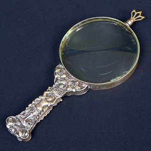 Lupa com cabo em metal ricamente trabalhado, apresentando figura feminina entre guirlandas e flores. Anéis com arabescos em relevo contornam a moldura da lente. Compr. 18,5 cm