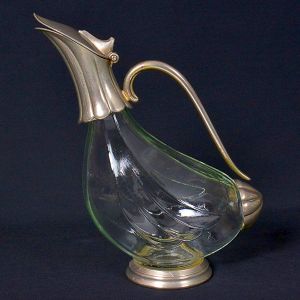 <b>Garrafa </b>em vidro lapidado com montagem em metal prateado. Corpo de linhas curvas, decorado por entalhes discretos, contrastando com bico e alça em metal trabalhado. Alt. 26 cm