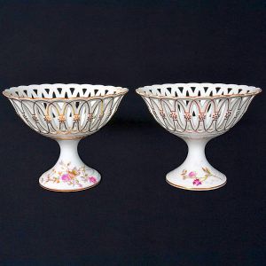 <b>Par de fruteiras de pé alto</b>  em porcelana francesa Limoges, decoradas com esmaltes policromados em padrões florais e frisos dourados. Bordas recortadas. Alt. 18 cm; Diâm. 23 cm