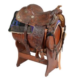 <b>Sela </b>tradicional em couro ricamente trabalhado com ferragens decorativas, apoiada sobre barril de madeira. Acompanha pelego e mantas em tecido colorido. Alt. total 104 cm; Compr. da sela 69 cm