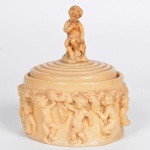 <b>Pote com tampa</b>, em marfim esculpido, decorado com figuras de "puttis" em relevo ao redor do corpo e figura de menino sentado sobre a tampa. Alt. 14 cm