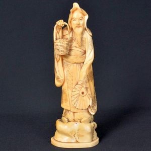 <b>Escultura em marfim</b>  representando monge ou sábio chinês, segurando cesta e leque. Detalhes intrincados na vestimenta e na barba da figura. Alt. 20 cm