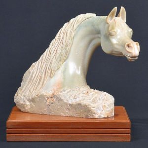 <b>Escultura </b>representando cabeça de cavalo, em pedra natural, cinzelada em bloco irregular, com base em madeira. Assinada Tradee na lateral. Alt. 32 cm