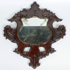<b>Espelho </b>de moldura entalhada em madeira escura, de inspiração rococó, trabalhada com volutas e concheados assimétricos. Alt. 88 cm