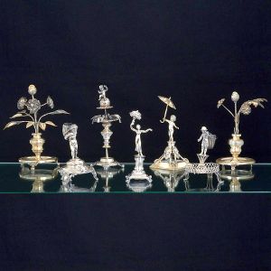 <B>Coleção de 7 paliteiros</B>, em prata portuguesa: 1) Bebedouro com aves encimado por figura infantil apoiada em globo e pés de garras. Alt. 22 cm; 2) Figura de dançarina sustentando cornucópia de flores e apoiada em base cilíndrica sobre "plateau" e pés em garras; 3) Figura angelical com guarda-chuva apoiada em base com carrancas antropomorfas entre espadas; 4) Figura masculina com cesto sobre base ovoide e vazada; 5 e 6) Em prata brasileira. Decorados com vasos de flores sobre base circular guilhochada. Alt. 22 cm; 7) Figura infantil sustentando vaso às costas (vestígio de contraste). Peso 1920 gramas.