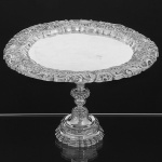 Excepcional <b>esmoleira </b>de coleção em prata brasileira do séc. XVIII. Centro liso. Borda e suporte em magnífico trabalho de repuxados e cinzelados com os elementos característicos do período barroco, onde se destacam as elaboradas composições de motivos vegetais com flores, volutas, folhas de acanto e concheados. A haste destacável apresenta elegante movimento apresentando os mesmos elementos acrescidos de folhas de palma, pequenos botões, frisos e perolados. Aparentemente sem marcas. Alt. 25 cm; Diâm. 38,5 cm. Peso 1435 gramas.