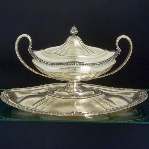 <b>Sopeira com seu "presentoir" </b>em prata portuguesa 1º título. A sopeira assenta-se em "plateau" levemente elevado ao centro e possui forma navicular com alças recurvadas. Decoração das bordas em godrões intercalados por folhas de acanto e pega da tampa modelada em pinha. A peça pode ser reversível também para floreira, pois acompanha placa interna destacável em composições vazadas. Contraste da cidade do Porto usado a partir de 1938. Compr. do "presentoir" 60,5 cm; Compr. da sopeira 48 cm. Peso 6120 gramas.