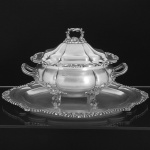 <B>Sopeira </B>em prata inglesa de forma oblonga. Apresenta bojo e tampa gomados, pega e pés em repuxados e cinzelados com elementos fitomorfos e "coquilles". Contraste da cidade de Sheffield de 1909 e marca do prateiro Walker & Hall. Acompanha "presentoir" do período George III, de borda ondulada com decoração muito similar. Contraste da cidade de Londres de 1796 e marca de prateiro. Compr. Da sopeira 37,5 cm; Compr. do "presentoir" 54 cm. Peso 5010 gramas.