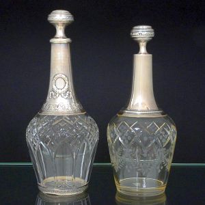 <b>Conjunto de duas garrafas </b>francesas em cristal com gargalo e tampa revestidas em prata. Uma delas com guirlandas e motivos de águias intercaladas por estilizações. A outra com lapidação em guirlandas, elementos geométricos e filetado de elementos circulares. A primeira com contraste do séc. XIX e vestígios de marca de prateiro. A segunda com mesmo contraste e marca de prateiro E G. Alt. 27,5 cm e 25,5 cm