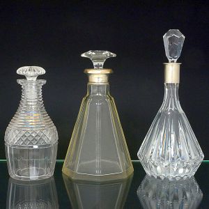 <b>Conjunto de 3 garrafas </b>em cristal transparente com lapidações distintas. Uma de corpo cilíndrico e bojo cônico, com lapidação em faixas diagonais e tampão facetado em formato abaulado, outra de corpo prismático com seis faces, gargalo com anel metálico e tampão em cristal facetado e a terceira, de corpo periforme, lapidação vertical em gomos longos terminada em tampão facetado de corte geométrico.