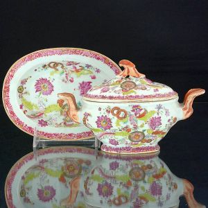 Rara <b>sopeirinha para molho e seu "presentoir" </b>em porcelana Companhia das Índias decorada em policromia nos esmaltes da Família Rosa numa das variações do padrão "Folha de Tabaco", conhecida como "Folha de Chá". Peça oblonga com alças e pega da tampa em "rouge de fer" realçado a ouro. Séc. XVIII. Desprezível bicadinho em uma das alças .Compr. do "presentoir" 19 cm; Compr. da terrina 21 cm