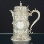 "<b>Flacon</b>" em prata inglesa vitoriana decorada com máscaras antropomorfas bacantes e elementos circulares sobre profusão de arabescos texturizados. Bico saliente. Pega da tampa abobadada com pega em pião invertido. Alça recurvada com isoladores de marfim. Interior em "vermeil". Contraste da cidade de Londres de 1876 e marca ilegível de prateiro. Alt. 27 cm. Peso 1140 gramas.