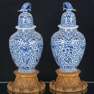 Belíssimo <b>par de potiches </b>em porcelana holandesa de Delft, decorados com arabescos floridos em azul índigo sobre fundo branco, ao gosto das porcelanas Companhia das Índias do período Kangxi. Pega da tampa modelada em pássaro e base em madeira entalhada com vestígios de douração. Porcelana com assinatura no verso. Alt. sem a base 46,5 cm; Alt. com a base 58,5 cm 