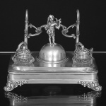 <b>Tinteiro </b>em prata apresentando bandeja retangular com dois recipientes para tintas com bojo de cristal e ao centro sineta encimada por figura angelical com lira. Suporte posterior com galhos de parreira e cachos de uvas. Inscrições na base e na sineta. Datada de 15/05/1904 (minúscula falta na ponta de 1 galho à esquerda). Med. 16 x 22 x 16 cm