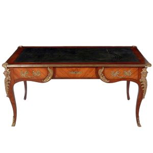 Belíssimo "<b>bureau plat</b>" francês, Régence, do séc. XVIII, construído em madeira europeia "rosewood". Caixa retangular com abas laterais e forração em couro negro pirogravado. Três gavetas anteriores e pernas recurvadas. Ricas ferragens protetoras e guarnições com cabeças femininas emplumadas. Med. 76 x 157 x 87 cm