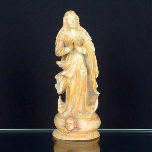 Imagem de <b>Nossa Senhora</b>, esculpida em marfim. A santa apresenta-se de pé, com mãos postas em oração, trajando manto esvoaçante com pregas bem definidas e ornamentos rendilhados. Base circular decorada, apresentando na parte frontal uma cabeça de querubim, elemento típico da iconografia barroca. Alt. 17,5 cm