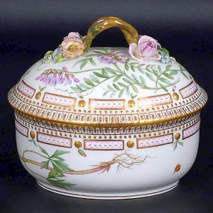 <b>"Bombonnière" com tampa </b>em porcelana dinamarquesa da importante manufatura Royal Copenhagen modelo "Flora Danica". Peça inteiramente pintada à mão em belíssima policromia com suas designações botânicas e numeração no verso. Apresenta borda em mínimos picotados cobertos a folha de ouro. Compr. 15,5 cm