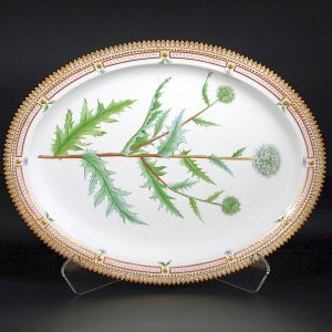 Grande <b>travessa </b>oval em porcelana dinamarquesa da importante manufatura Royal Copenhagen modelo "Flora Danica". Inteiramente pintada à mão em belíssima policromia com suas designações botânicas e numeração no verso. Apresenta borda em mínimos picotados cobertos a folha de ouro. Med. 47 x 36,5 cm