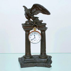 <b>Relógio de mesa </b>em bronze, simulando ruínas clássicas com duas colunas caneladas sustentando um entablamento. Na parte superior, figura de águia alçada. Ao centro, pendurado sob o arco, relógio de bolso adaptado como mostrador, com numeração arábica e visor branco. Alt. 23 cm