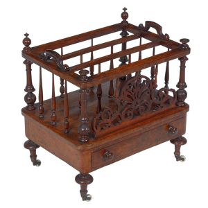 Belo <b>porta-revista </b>"Canterbury" inglês, em raiz de nogueira. Três divisórias com montantes em balaústre e alças laterais. Parte anterior com entalhe de arabescos e gaveta à frente. Pernas curtas torneadas terminadas em rodinhas de época. Séc. XIX. Med. 59 x 56 x 42 cm