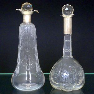<b>Par de garrafas </b>em cristal com delicado trabalho de lapidação geométrica e floral, apresentando tampas facetadas montadas em gola de prata inglesa. Uma peça possui corpo afilado com desenho vertical em espiral, a outra, bojo arredondado com lapidações radiantes.