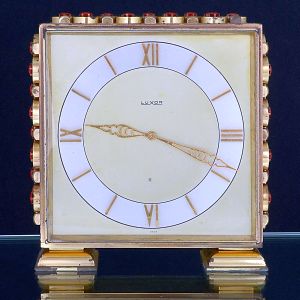 <b>Relógio de mesa Luxor</b>, com caixa quadrada, em metal dourado decorado nas laterais com pedras circulares em tom rubi. Mostrador com algarismos romanos estilizados e ponteiros trabalhados em metal dourado. Base igualmente em metal dourado, com desenho escalonado.