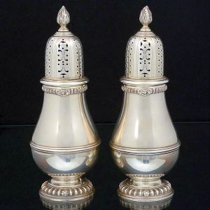 <b>Par de polvilhadores </b>em prata inglesa lisa piriformes e pesados. Decorados com frisos na parte bojuda e gomos intercalados por folhas na borda. Pegas das tampas em modelados de pinha. Contraste da cidade de Londres de 1960. Prateiro G & C LD. Alt. 20,5 cm