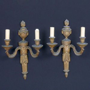 <b>Par de apliques de parede</b>, em estilo neoclássico, executados em bronze dourado. Corpo central em forma de urna encimada por chama, ladeado por dois braços em voluta, ricamente ornados, sustentando "bobèches" e simulando velas. Alt. 45 cm