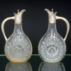 <b>Par de jarras </b>inglesas para vinho em cristal lapidado com motivos geometrizados. Gargalo e alça em prata vitoriana decorada com laços e guirlandas floridas. Contraste da cidade de Sheffield de 1893. Marca do prateiro H H. Alt. 25,5 cm