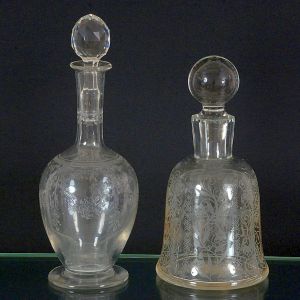 <b>Par de garrafas </b>em cristal transparente decorado com finos motivos florais e arabescos gravados. Uma apresenta bojo arredondado e tampa facetada, a outra, corpo em formato acampanado e tampa esférica lisa.