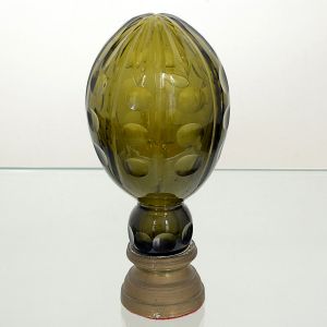 <B>Pinha </B>decorativa em vidro verde, com superfície trabalhada em relevos ovais dispostos em linhas verticais. Corpo de forma alongada, assentado sobre base em latão de perfil escalonado. Alt. 19 cm