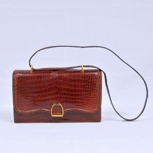 HERMÈS - Paris - Antiga <b>bolsa </b>em couro de crocodilo na cor marrom com ferragens douradas, tendo o fecho forma de estribo. Med. 26 x 17 x 6,5
