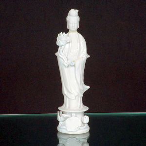 <b>Escultura </b>em porcelana "blanc de chine", representando divindade de pé, segurando flor de lótus, sobre base ornamentada. Alt. 21 cm