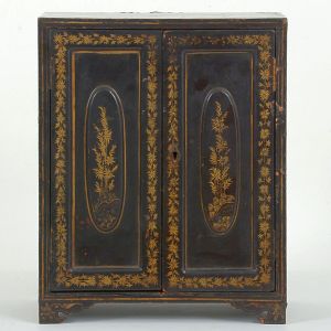 Pequeno <b>armário </b>chinês em madeira laqueada de negro, apresentando duas portas anteriores com reservas almofadadas de forma oval e interior com cinco gavetas com puxadores em botões circulares. Realces a ouro com ramagens de bambu e filetados. Início do séc. XIX ou fins do XVIII. Med. 35,5 x 28,5 x 17,5 cm