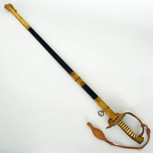 <b>Espada </b>de oficial, de gala, com bainha em madeira revestida e guarnições metálicas douradas, ricamente trabalhadas com motivos decorativos em relevo. Guarda do tipo cálice vazado, igualmente decorada. Na guarnição da bainha, inscrição "DOURADA M18" acompanhada de monograma.