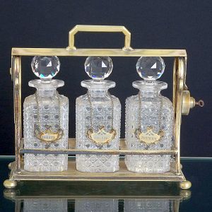 Antigo <b>"tantalus" </b>em metal inglês, apresentando gabinete com três "decanters" em cristal lapidado em "cabochons" e colares também em metal. Punção do fabricante no verso. Alt. 28,5 cm; Compr. 32,5 cm