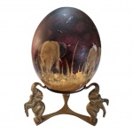 Ovo de avestruz pintado à mão com figuras representando elefantes, apoiado em base triplóide em bronze representando elefantes, medidas do ovo: 16 x 14 cm. base: 13 x 9 cm.