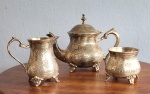 Três peças para servir café / chá em metal prateado, asiático, rico trabalho em baixo relevo sendo uma chaleira (17x20cm), uma leiteira (11x12cm) e um açucareiro (10x8cm)..
