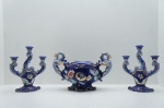 Suntuoso Jogo Trio em Porcelana Zappi (Brasil, meados do Século XX), composto por um vaso potiche central e um par de castiçais de três braços. As peças apresentam um vibrante azul cobalto enriquecido por decoração floral policromada e profusos adornos em ouro. O vaso destaca-se pelas alças em forma de dragões, conferindo um estilo opulento. Marcado com "Zappi" na base. Ideal para colecionadores de porcelana brasileira de luxo. Centro de mesa com trincado colado no fundo. Centro de mesa: 27 x 41 x 20 cm. / Candelabro alt. 31 cm.